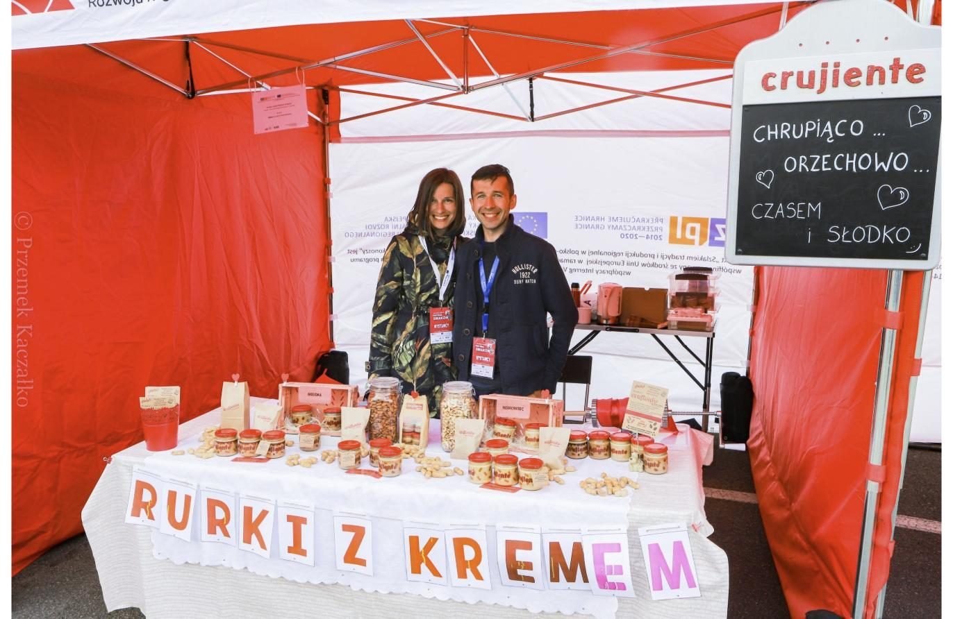 Magdalena i Paweł za stołem pełnym słoików na targu, nad nimi tablica Crujiente: chrupiąco, orzechowo, czasem i słodko